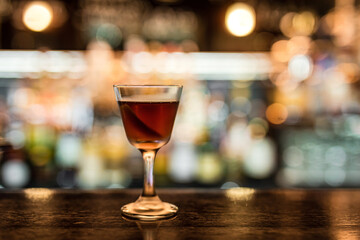 Wall Mural - A glass with cold Manhattan cocktail garnished with orange zest peel on a wooden bar counter. Bokeh lights, selective focus, small depth of field