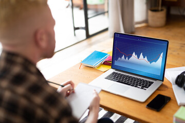 Poster - Over shoulder view of caucasian businessman writing notes and using laptop with graphs screen