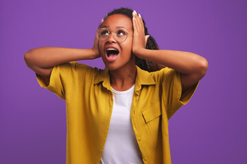 Wall Mural - Young stunned African American wow woman clutching head and screaming in surprise or delight at what she sees above dressed in casual clothes stands in purple studio. Emotional ethnic girl