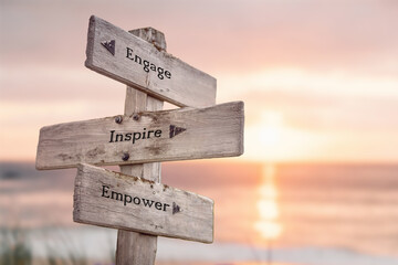 engage inspire empower text quote engraved on wooden signpost outdoors on the beach with sunset theme.