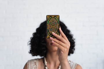 Wall Mural - KYIV, UKRAINE - JUNE 29, 2022: young brunette oracle obscuring face with tarot card during spiritual session.