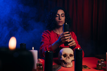 Wall Mural - oracle with closed eyes holding burning candle near skull and blue smoke on dark background.
