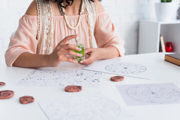 Wall Mural - partial view of fortune teller holding bottle of essential oil near blurred clay runes and natal charts.