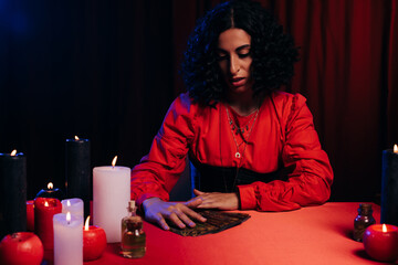 Wall Mural - young brunette predictor near tarot cards and burning candles on red table and dark background.