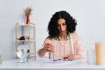 brunette astrologer drawing star charts near crystal ball at home.