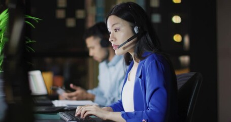 Wall Mural - video of asian businesswoman talking on headset, working at night with colleagues in office