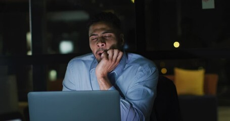Wall Mural - video of tired biracial businessman sitting at desk using laptop, working at night in office