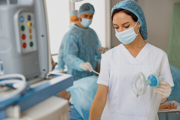 Wall Mural - Nurse holding breathing mask for anesthesia standing in operation room with colleagues on background