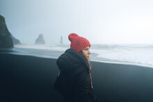 A Woman In A Black Jacket And Winter Hat Is Standing On A Black Beach In Iceland. Hazy Rocks And Waves In The Background. Misty Rainy Day.