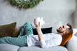 Thoughtful bearded man lying on a sofa holding white piggy bank. Man making a wish, dreaming of money. Christmas expenses, a way to get rich concept.  Selective focus.