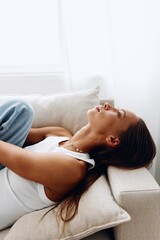 Wall Mural - A beautiful young woman lies at home on the couch in a relaxed pose in a white T-shirt and blue jeans. Lifestyle in comfort