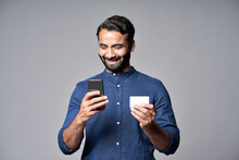 Happy Indian Business Man Holding Smartphone And Credit Card Using Cell Phone Mobile Bank App Making Convenient Financial E Commerce Payment Digital Transaction Paying Online Isolated On Gray.