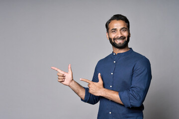 Happy indian business man salesman looking pointing fingers hand gesture aside indicating advertising recommending new commercial promotion, presenting sale offer standing isolated on gray background.