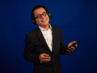 Portrait of smart middle-aged asian confident successful businessman wearing suit listening music with wireless headphones and smartphone on blue background in studio, looking happy leadership