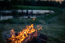 Leaping Flames Of A Camp Fire With A Darkening View Of A Pond In The Background.