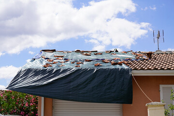 Storm damaged roof on house with a black plastic tarpaulin over hole in the shingles and rooftop after spring summer thunderstorm violent