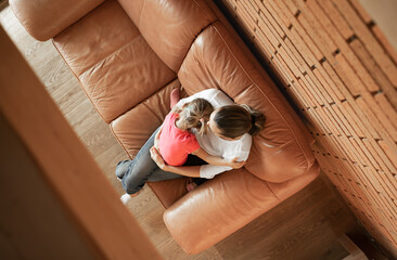 Wall Mural - Top view of single mother sitting on the couch at home holding her child daughter. Motherhood, and parenting concept. Tired stay at home mother taking a break form busy day. 
