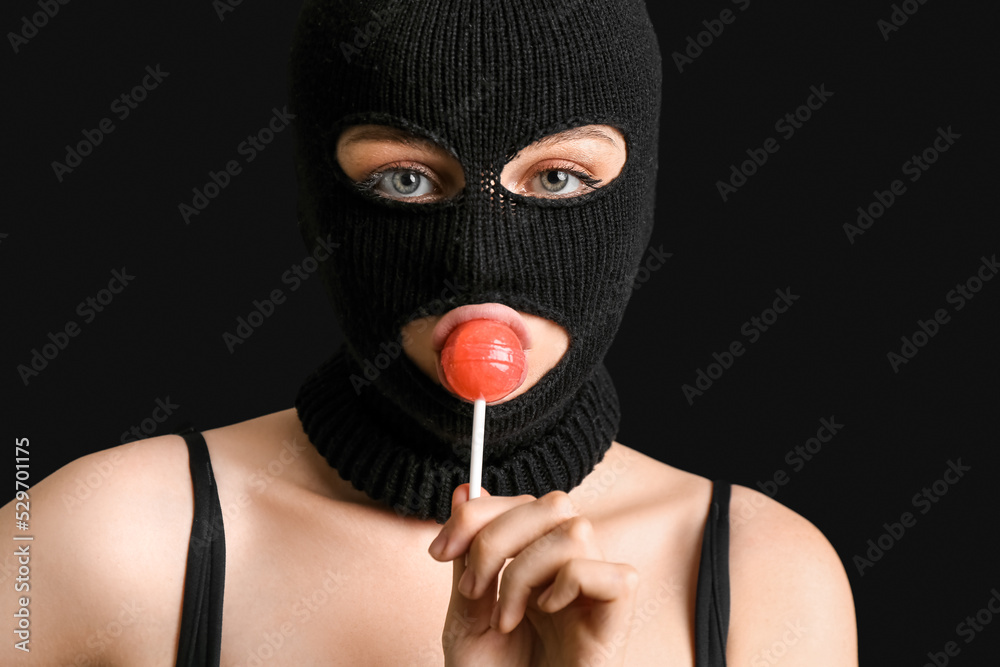 Young woman in balaclava with lollipop on black background