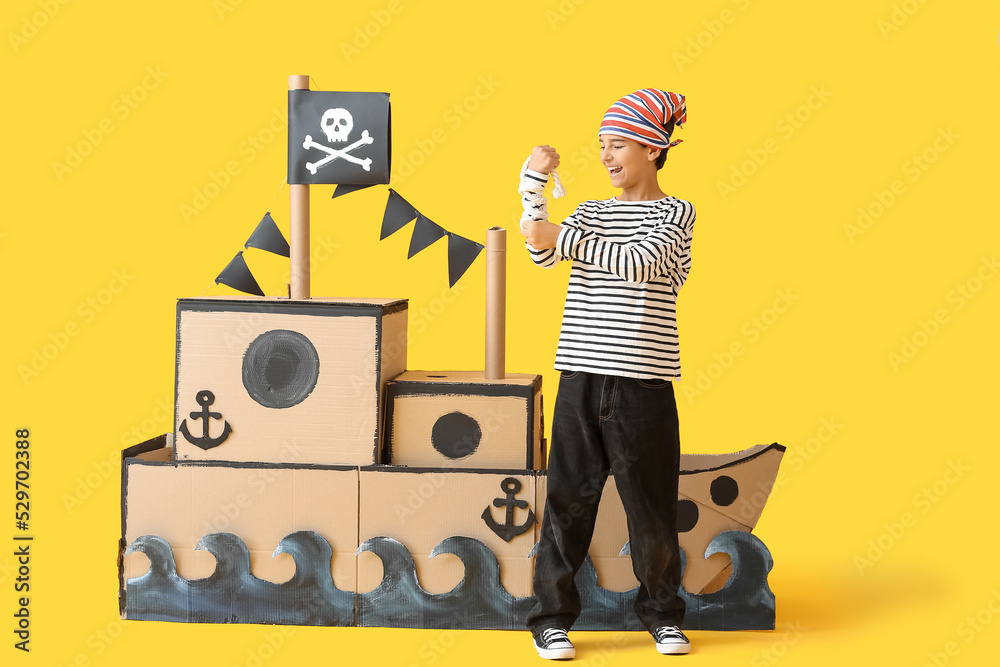 Little boy dressed as pirate with rope and cardboard ship on yellow background