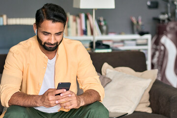 Wall Mural - Happy indian man user sitting at home relaxing on couch wearing earbuds using cell phone looking at smartphone watching social media videos, listening music podcast, having virtual mobile chat call.