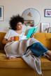 © Daniel - Relaxed, happy African american woman at home living room sitting on the couch, reading a book and drinking tea