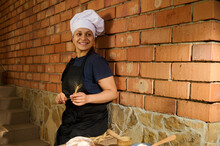 Portrait Of A Smiling Pretty Woman, A Baker Confectioner Posing With Spikelet Of Wheat Against Red Brick Wall Background