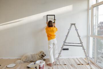 Wall Mural - Young woman hanging picture frame in room, decorating her newly renovated apartment, stands with her dog in white room