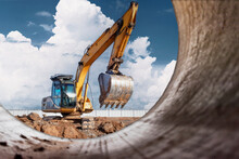 A Powerful Caterpillar Excavator Digs The Ground Against The Blue Sky. Earthworks With Heavy Equipment At The Construction Site. View From A Large Concrete Pipe.