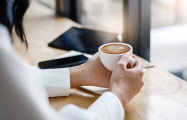 Close up shot young asian woman holding coffee cup with latte coffee she sitting at counter in cafe with mobile, digital tablet and note book placed nearby. she relaxing after work