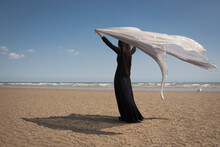 Fine Art Beach Scene With Woman In Long Black Dress Holding Thin Fabric In The Wind