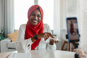 Happy african american islamic blogger showing skincare product to cellphone camera, making video for her beauty blog