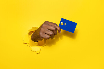 Wall Mural - Banking services. African american male hand holding plastic credit card through the torn yellow studio background