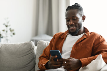 Aufkleber - Cheerful african american man using new app on cellphone and smiling, sitting on sofa at home, free space