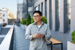 © Prostock-studio - Positive asian middle aged male entrepreneur checking emails on smartphone while walking by business center