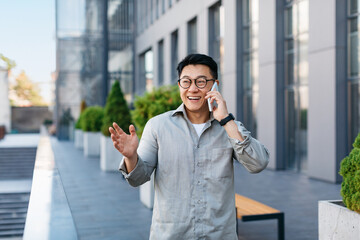 Happy successful asian businessman talking on smartphone, standing outdoors against modern office center, free space