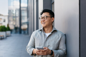 Confident korean middle aged businessman holding smartphone and looking aside at free space, standing outdoors