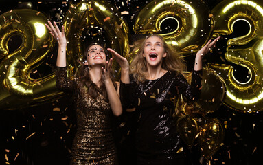 Happy gorgeous girls in stylish sexy party dresses holding gold 2023 balloons, having fun at New Year's Eve Party.