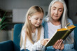 © Prostock-studio - Serious caucasian small granddaughter and mature grandmother are reading book on sofa in room interior