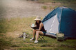 Happy woman sitting on the camping chair drinking coffee and reading a book in front of the camping tent at meadow near lake. Recreation and journey outdoor activity lifestyle.