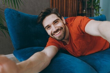 Wall Mural - Close up young smiling man wear red t-shirt do selfie shot pov on mobile cell phone lay down blue sofa stay at home hotel flat rest spend free spare time in living room indoors People lounge concept.