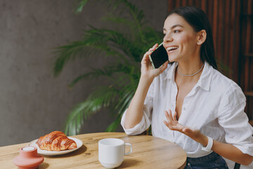 Young fun smiling calm cheerful happy latin woman 30s wearing white shirt talk speak on mobile cell phone sit alone at table in coffee shop cafe restaurant indoors. Freelance office business concept.