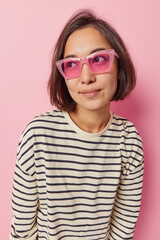 Wall Mural - Lovely young dark haired Asian woman looks away thoughtfully daydreams about something wears trendy sunglasses and casual striped jumper isolated over pink background. People and thoughts concept