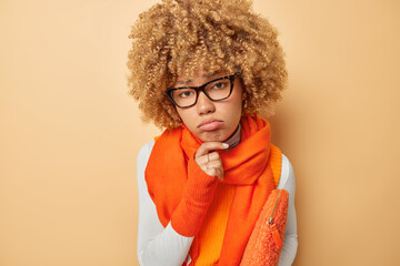 Wall Mural - Portrait of distressed upset female model keeps hand on chin feels frustrated wears spectacles orange scarf around neck carries small bag under arm looks sadly at camera isolated over beige wall