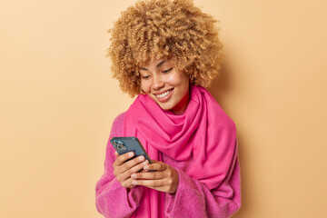 Wall Mural - Positive good looking young woman with curly hair receives pleasant notification watches video via smartphone wears pink jumper and scarf isolated over beige background communicates in internet