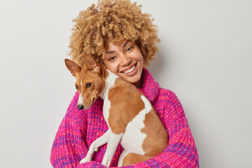 Carefree positive curly woman hugs basenji dog takes care of cute pet enjoy spending weekend together dressed in pink knitted sweater isolated over grey background. People and animals concept