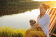 © sementsova321 - Side view portrait of happy positive woman wearing white shirt sitting with her Pekingese dog near the river on deck chair and talking on mobile phone, having pleasant conversation.