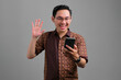 Portrait of smiling young Asian man wearing batik shirt saying hi and waving hand while making video call on smartphone isolated on grey background