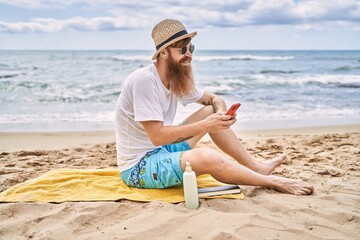 Sticker - Young redhead man using smartphone sitting on the towel at the beach.