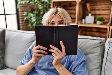 Poster - Young blond man covering mouth with book sitting on sofa at home