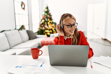 Poster - Young caucasian girl sitting on the table working using laptop by christmas tree smiling with hand over ear listening an hearing to rumor or gossip. deafness concept.